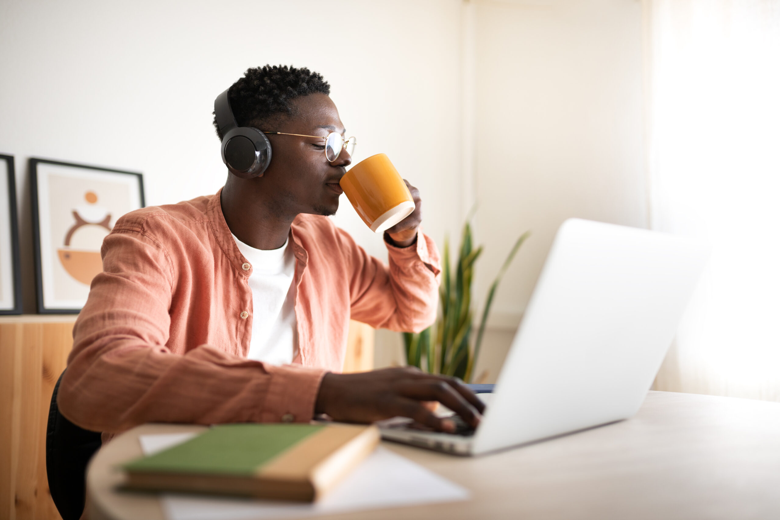 Young African American black man drinking coffee while listening to music and working with laptop. Working from home. E-learning.